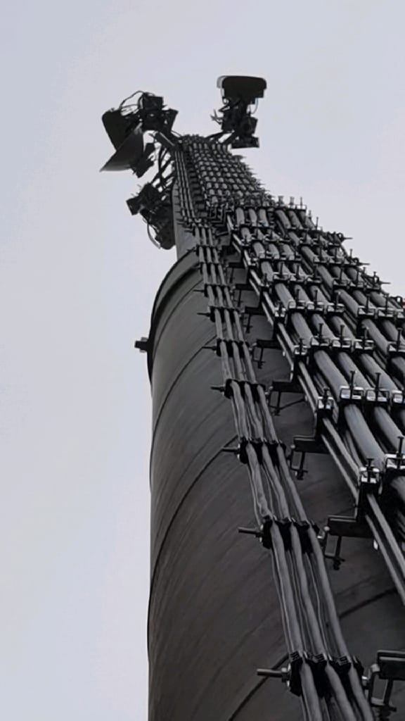 An upward view of a tall mast carrying antennas and cables.